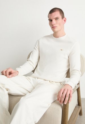 Crème-kleurig overhemd met lange mouwen in textuur katoen met een ronde halslijn, voorzien van een klein logopatch op de borst. Het model draagt bijpassende broek.
