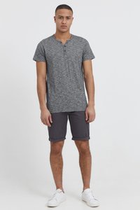 Grå melange kortærmet henley skjorte med tre knapper, parret med oprullede mørke shorts og hvide sneakers, stående foran en ensfarvet baggrund.