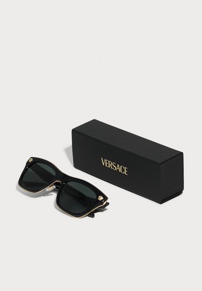 Black Versace sunglasses with oversized square frames, gold-accented edges, and dark lenses, placed next to a black box with gold logo.