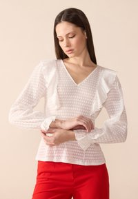Hvit strukturert bluse med volangermidler og V-hals. Har et subtilt mønster og et tettsittende design, kombinert med røde bukser.