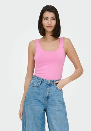 Woman with straight dark hair wears pink sleeveless top and high-waist blue jeans, standing with one hand in pocket against light background.