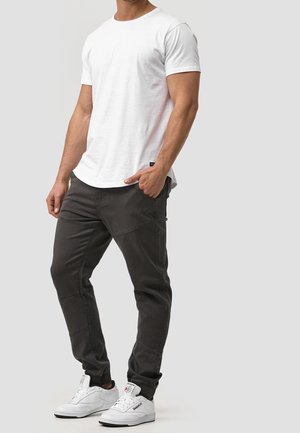 Homme portant un t-shirt blanc à manches courtes, un pantalon de jogging gris foncé avec des poches et des baskets Reebok blanches, se tenant devant un fond clair.