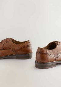 Chaussures en cuir marron avec un design perforé, des détails en forme d'aile et un petit talon empilé. Lacets et coutures contrastantes visibles.