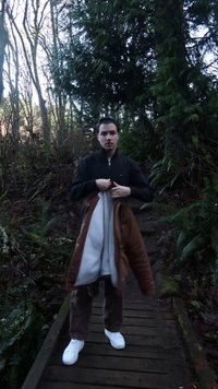 Bruine jas over een grijze trui, gecombineerd met een bruine broek en witte sneakers. Omringd door groen op een houten pad.