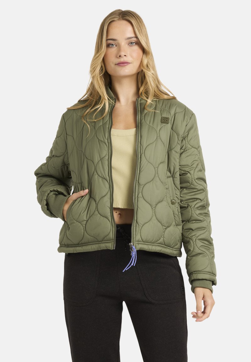 Blond kvinde iført olivengrøn quilted jakke over en beige cropped top og sorte bukser, med den ene hånd i jakke-lommen.