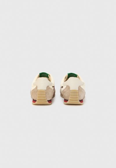 Μπεζ σουέτ και κρεμ δερμάτινα αθλητικά παπούτσια με υφή στη φτέρνα και πράσινες λεπτομέρειες. Διαθέτουν κόκκινη μεσαία σόλα και σχεδιασμό ριγωτής εξωτερικής σόλας.