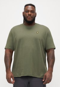 Tricou scurt din bumbac moale, de culoare verde măslin, cu mânecă scurtă, guler rotund și un mic logo brodat galben pe piept. Croială obișnuită.