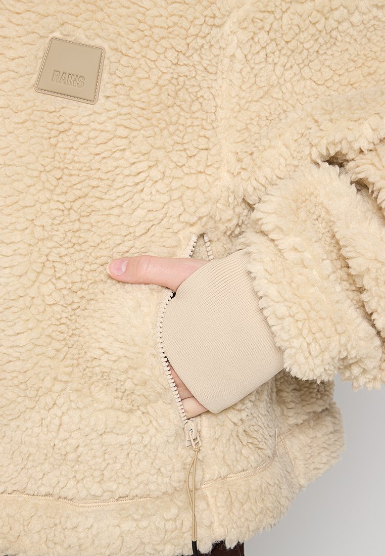 Chaqueta de piel sintética beige con una superficie suave y texturizada. Presenta un bolsillo con cremallera lateral y puños acanalados. Parche con el logo en la parte delantera.