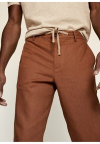 Pantalons en lin marron avec une taille à cordon, coupe droite, poches avant et texture subtile, associés à un haut à manches courtes beige clair.