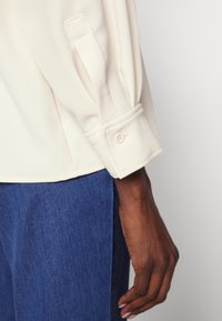 Cream blouse with a cuffed sleeve featuring a button detail, paired with blue denim. The fabric appears soft with a smooth texture.