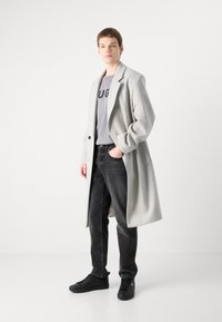 Abrigo de lana gris claro con solapa, usado sobre una camiseta gris con texto en negrita. Jeans negros y zapatillas completan el atuendo casual.