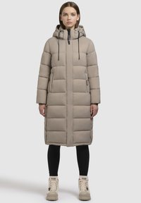 Langer, beiger Daunenmantel mit Kapuze, ausgestattet mit einem Reißverschluss, elastischen Kordeln und seitlichen Taschen. Getragen über schwarzen Leggings und beigen Stiefeln.