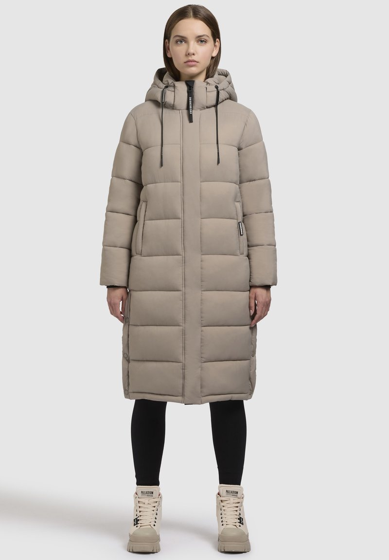 Langer, beiger Daunenmantel mit Kapuze, ausgestattet mit einem Reißverschluss, elastischen Kordeln und seitlichen Taschen. Getragen über schwarzen Leggings und beigen Stiefeln.