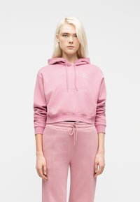 Νεαρός με μακριά ξανθά μαλλιά που φοράει ροζ κοντό φούτερ με κουκούλα και ροζ παντελόνι φόρμας με κορδόνια, κοιτάζοντας μπροστά.