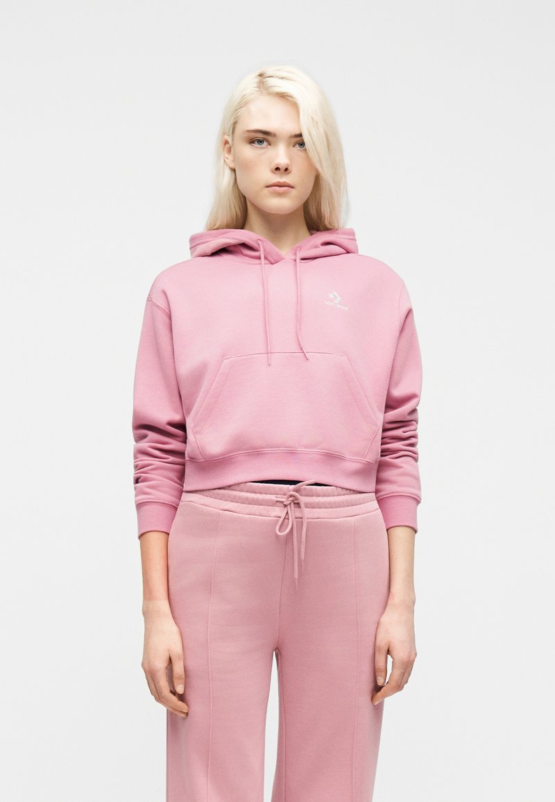 Νεαρός με μακριά ξανθά μαλλιά που φοράει ροζ κοντό φούτερ με κουκούλα και ροζ παντελόνι φόρμας με κορδόνια, κοιτάζοντας μπροστά.