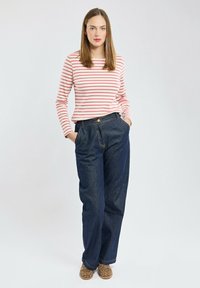 Maglia a maniche lunghe a righe rossa e bianca, abbinata a jeans a vita alta blu scuro. Calzature con stampa animale color brown sabbia. Modella in piedi con le mani in tasca.