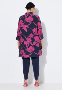 Floralbluse in Marineblau mit großen pinken Blumen, kombiniert mit eng anliegenden Marineblau-Hosen und pinken High Heels. Verfügt über einen Kragen und Dreiviertelärmel.