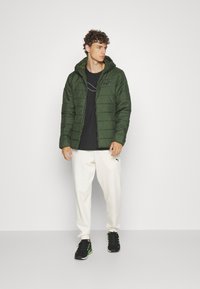 Giacca a vento verde con cappuccio, chiusura con zip e texture trapuntata; indossata su una maglietta nera, abbinata a pantaloni bianchi e sneaker nere.