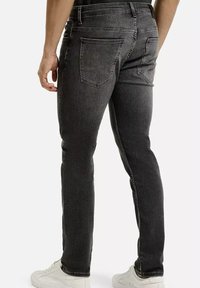 Mężczyzna w slim-fit wyblakłych czarnych dżinsach i białych sneakersach, stojący tyłem do kamery na jednolitym tle.