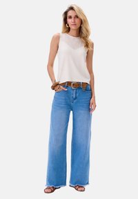 Witte mouwloze top met een gestructureerd bovenstuk, gecombineerd met lichtblauwe wijde jeans en een bruine riem. Bruine sandalen. Effen achtergrond.