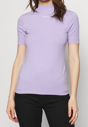 Kobieta ubrana w dopasowany jasnofioletowy top z krótkim rękawem z napisem "Calvin Klein Jeans" na piersi oraz czarne spodnie, stojąca na jasnym tle.