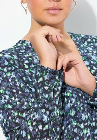 Multifarvet bluse med blomsterdesign i blå, grøn og sort, med V-udskæring og knappede ærmer, lavet af et let materiale.