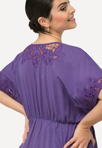 Violetti mekko, jossa on pitsidetelit olkapäillä ja hihoissa, virtaava kangas, kokoontuva vyötärö ja pehmeä rakenne, jossa on hienostuneita brodeerauksia.