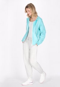 Giacca a vento blu chiaro con lacci bianchi, abbinata a pantaloni da jogging grigi con texture. Modello che indossa sneakers bianche con dettagli rosa.