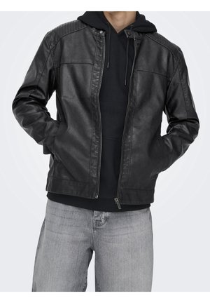 Homme portant une veste en cuir noire par-dessus un sweat à capuche noir et un jean gris clair, les mains dans les poches de la veste, sur un fond uni.