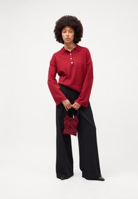 Suéter de polo tricotado em castanho-avermelhado com três botões, combinado com calças largas pretas. A segurar uma pequena mala vermelha texturizada.