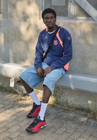 Marineblå og orange sportstrøje med logo, lyseblå denimshorts, hvide sokker, sorte og røde sneakers, siddende på en bænk.