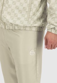 Helle beige Jacke mit kariertem Muster und elastischen Bündchen, kombiniert mit passenden beigen Hosen mit einem weißen Logo und glatter Textur.