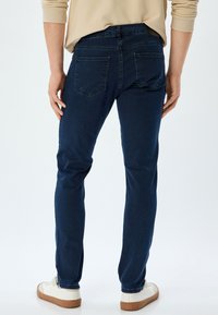 Jeans de denim azul oscuro con corte ajustado, que cuentan con dos bolsillos traseros y una textura suave, combinados con zapatillas blancas y una blusa beige.