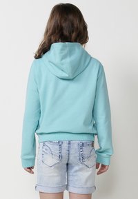 Tirkīza krāsas kapucis, kuram ir kangaru kabata, saskaņots ar gaiši zilām denim šortiem. Mīksta tekstūra, ikdienišķa piegriezuma, ar sarullētām apakšmalām šortiem.