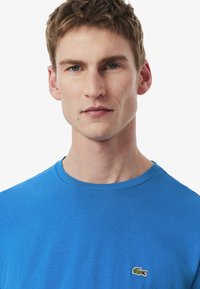 Camiseta de algodón azul con cuello redondo y logo de cocodrilo bordado en el lado izquierdo del pecho. Textura suave, diseño ajustado.