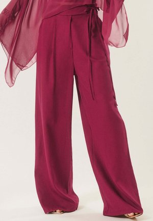 Femme portant un pantalon taille haute, jambes larges, de couleur magenta foncé, avec un haut assorti transparent tombant sur les hanches, debout sur un fond clair.