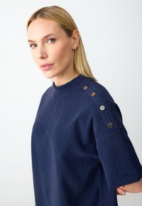 Navyblaues, strukturiertes Oberteil mit hohem Halsausschnitt und kurzen Ärmeln. Mit dekorativen metallischen Knöpfen an der Schulter für zusätzlichen Details.