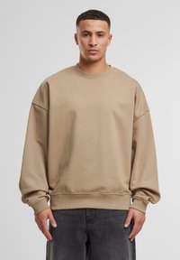 Overdimensioneret beige sweatshirt lavet af bomuld, med faldne skuldre, ribbet rund halsudskæring og manchetter, parret med mørkegrå jeans.