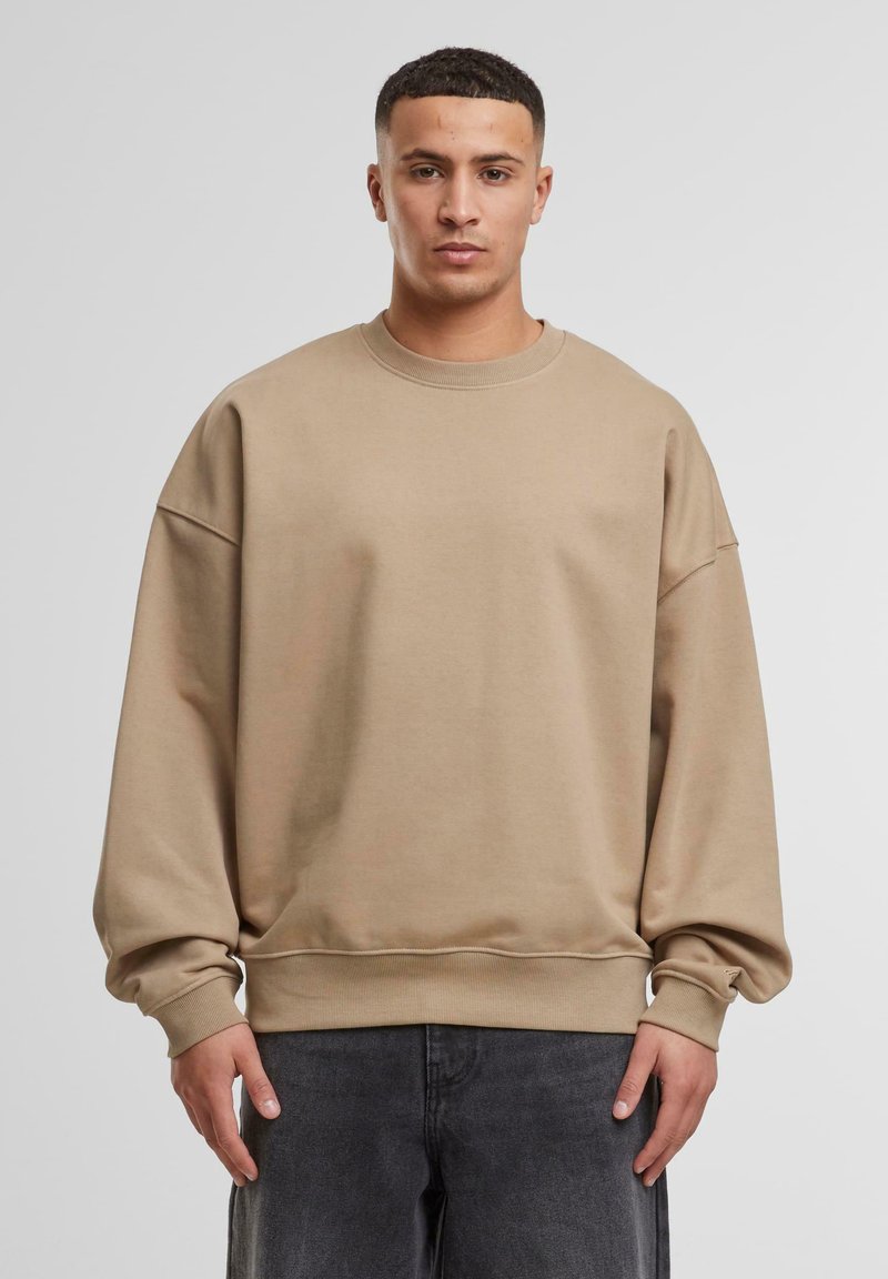 Overdimensioneret beige sweatshirt lavet af bomuld, med faldne skuldre, ribbet rund halsudskæring og manchetter, parret med mørkegrå jeans.