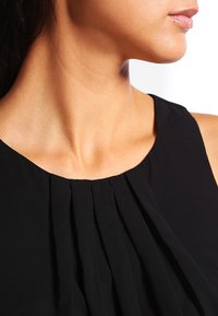 De nek en onderkant van het gezicht van een vrouw met een gladde huid, gekleed in een mouwloze zwarte top met verticale plooien bij de halslijn.
