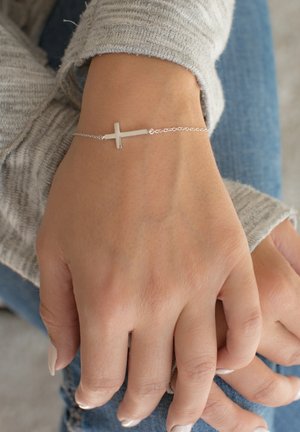 Egy ezüst láncos karkötő, amelyen egy egyszerű kereszt charm található, egy csuklón pihenve. Puha textúrájú, minimális dizájnnal; egy szürke pulóver háttérében.