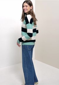 Kobieta uśmiechająca się, w pasiastym swetrze w kolorach teal, czerni i bieli, w połączeniu z szerokimi niebieskimi dżinsami, stojąca na tle o jednolitej barwie.