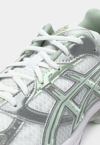 Λευκό αθλητικό παπούτσι με επάνω μέρος από δίχτυ και μεταλλικές ασημί και ανοιχτό πράσινες λεπτομέρειες, στρογγυλά κορδόνια και ενισχυμένη σόλα.