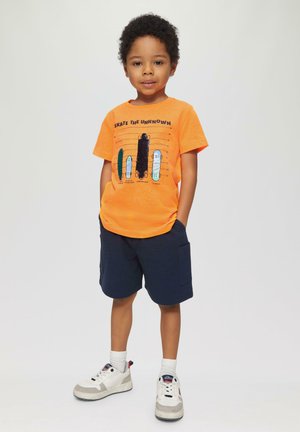 Felle oranje t-shirt met grafische print van skateboards, gecombineerd met marineblauwe shorts. Witte sneakers met grijze accenten en witte sokken.