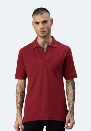 Uomo con capelli corti e tatuaggi sul collo e sulle braccia che indossa una polo rossa a maniche corte su uno sfondo chiaro e uniforme.