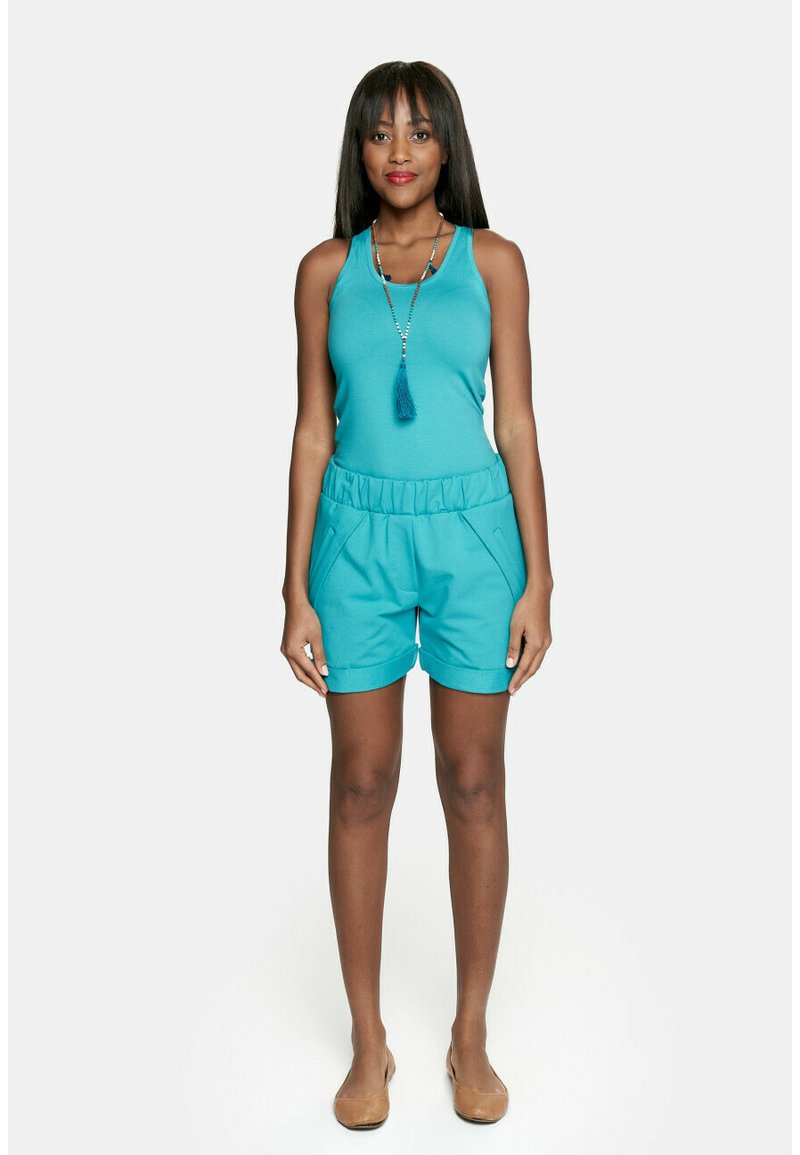Zielona bezrękawnikowa bluzka w połączeniu z odpowiadającymi szortami na elastycznej talii. Gładka tekstura, luźny krój, z dwiema bocznymi kieszeniami. Płaskie brązowe buty.