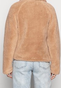Giacca in pelliccia ecologica di colore marrone chiaro, caratterizzata da una superficie morbida e testurizzata. Il design include maniche lunghe e un orlo arrotondato. Abbinata a jeans azzurri.
