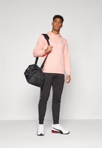 Lichtroze hoodie, zwarte sporttas, donkergrijze joggingsbroek, witte sneakers met rode accenten. Eenvoudig ontwerp, relaxte pasvorm en contrasterende kleuren.