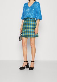 Top blu con paillettes e scollatura a V, abbinato a una mini gonna a quadri verde e nera. Scarpe con tacco quadrato nere completano l'outfit.