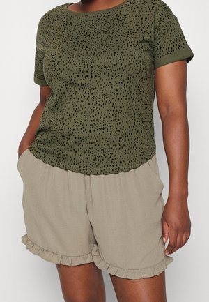 Kvinde iført en olivengrøn kortærmet skjorte med sorte pletter og beige shorts med flæsekant og hænderne i lommerne.
